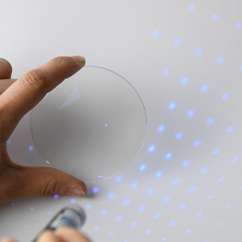 A blue cut lens held between two fingers under a violet torch, blue dots scattered around it